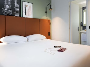 A hotel room with a bed, pillows, a framed picture of musical instruments, a nightstand with a lamp, and a bathroom visible through an open door.