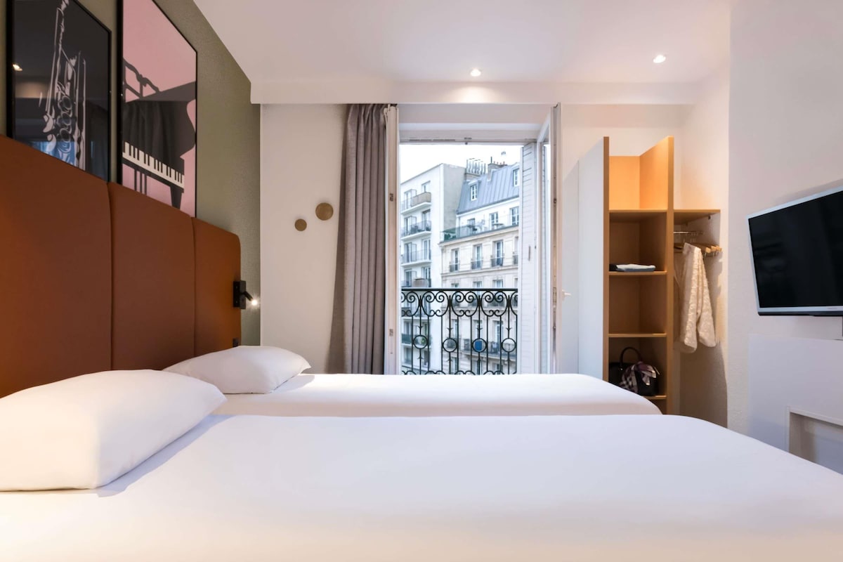 A hotel room with a large bed, a wardrobe, a television, and a balcony with a view of buildings.
