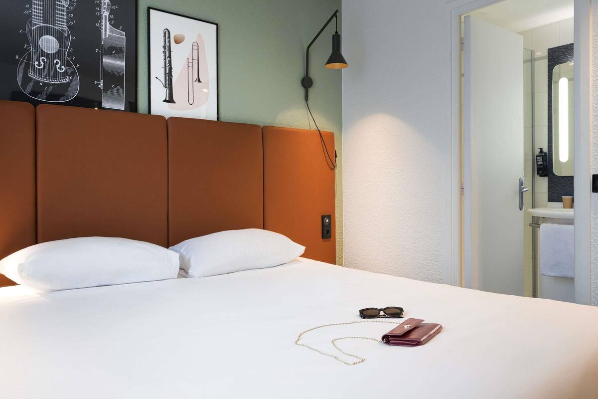 Een hotelkamer met een bed, kussens, een lijst met afbeelding van muziekinstrumenten, een nachtkastje met een lamp, en een badkamer zichtbaar door een open deur.