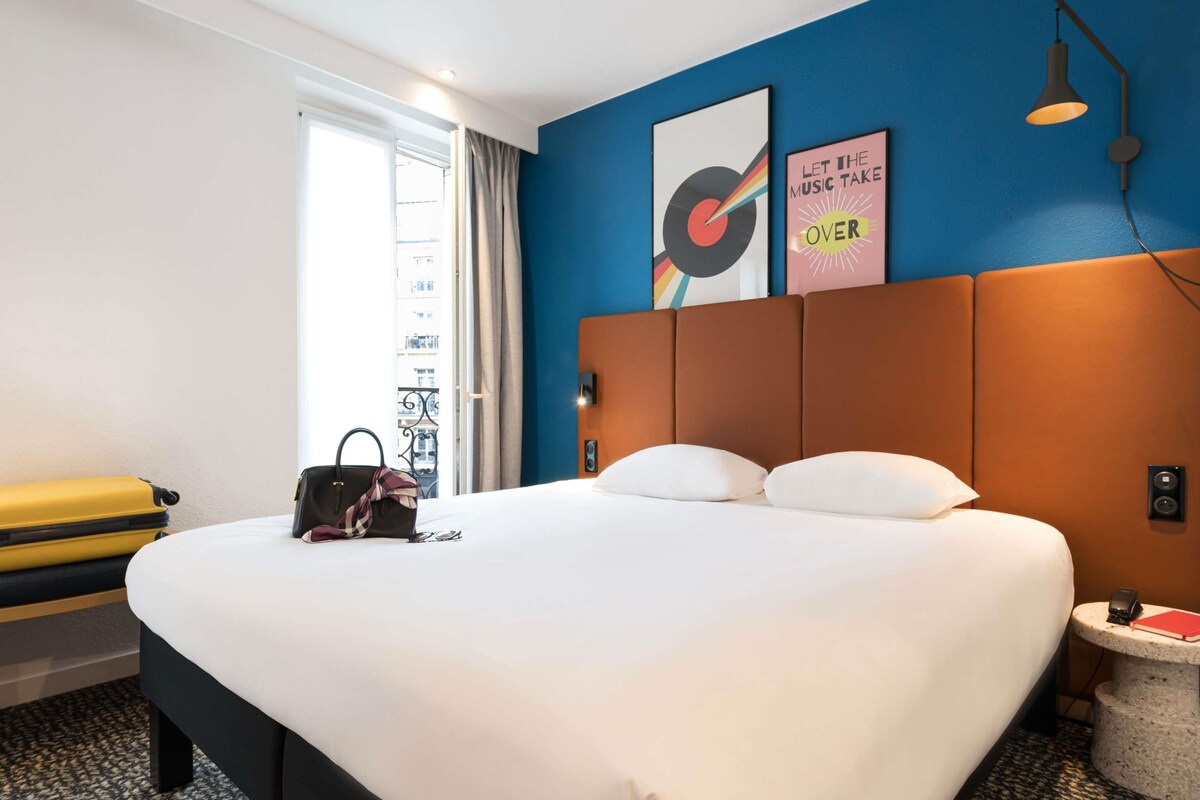 Een hotelkamer met een groot bed, een koffer, een handtas en een klein tafeltje met een boek.
