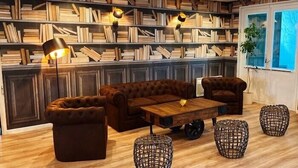 Un salon chaleureux avec un canapé en cuir marron, une table basse en bois et des poufs en osier. On y trouve des étagères remplies de livres et deux lampadaires.