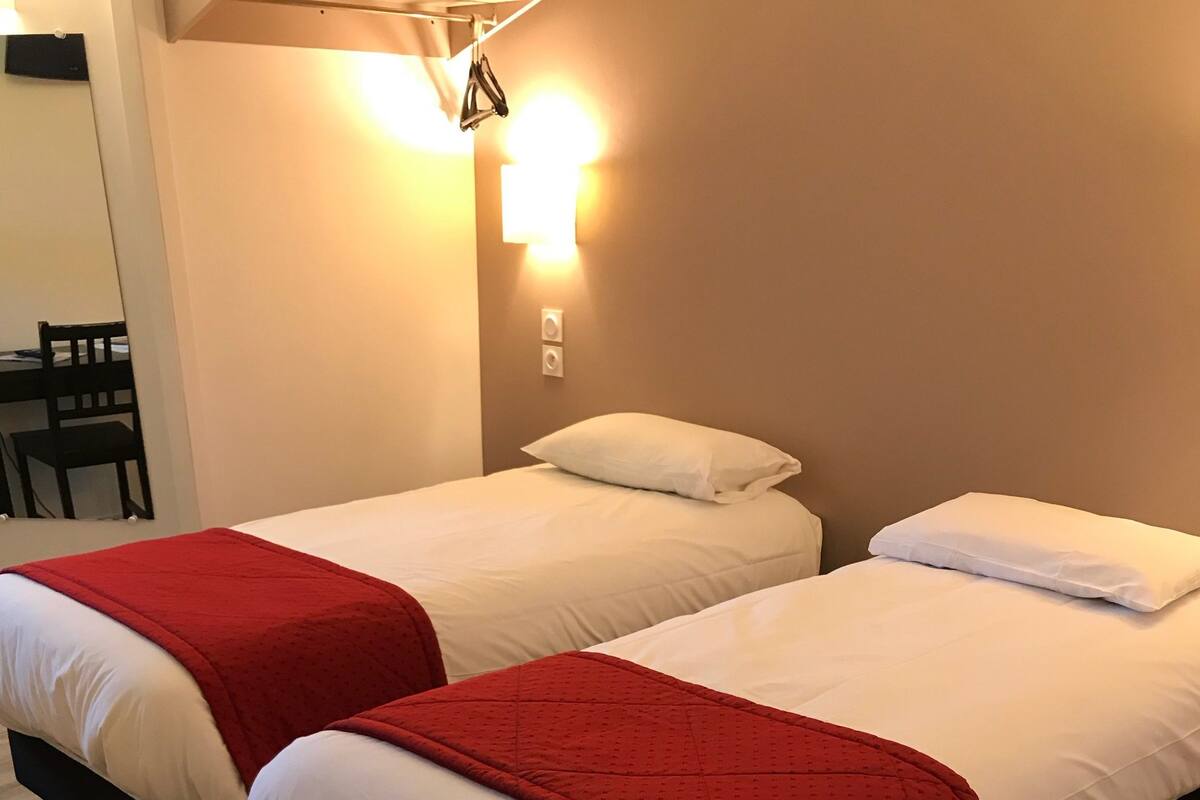 Two single beds with red blankets, wooden headboards, and white bedding in a room with wooden flooring and a wall-mounted light fixture.