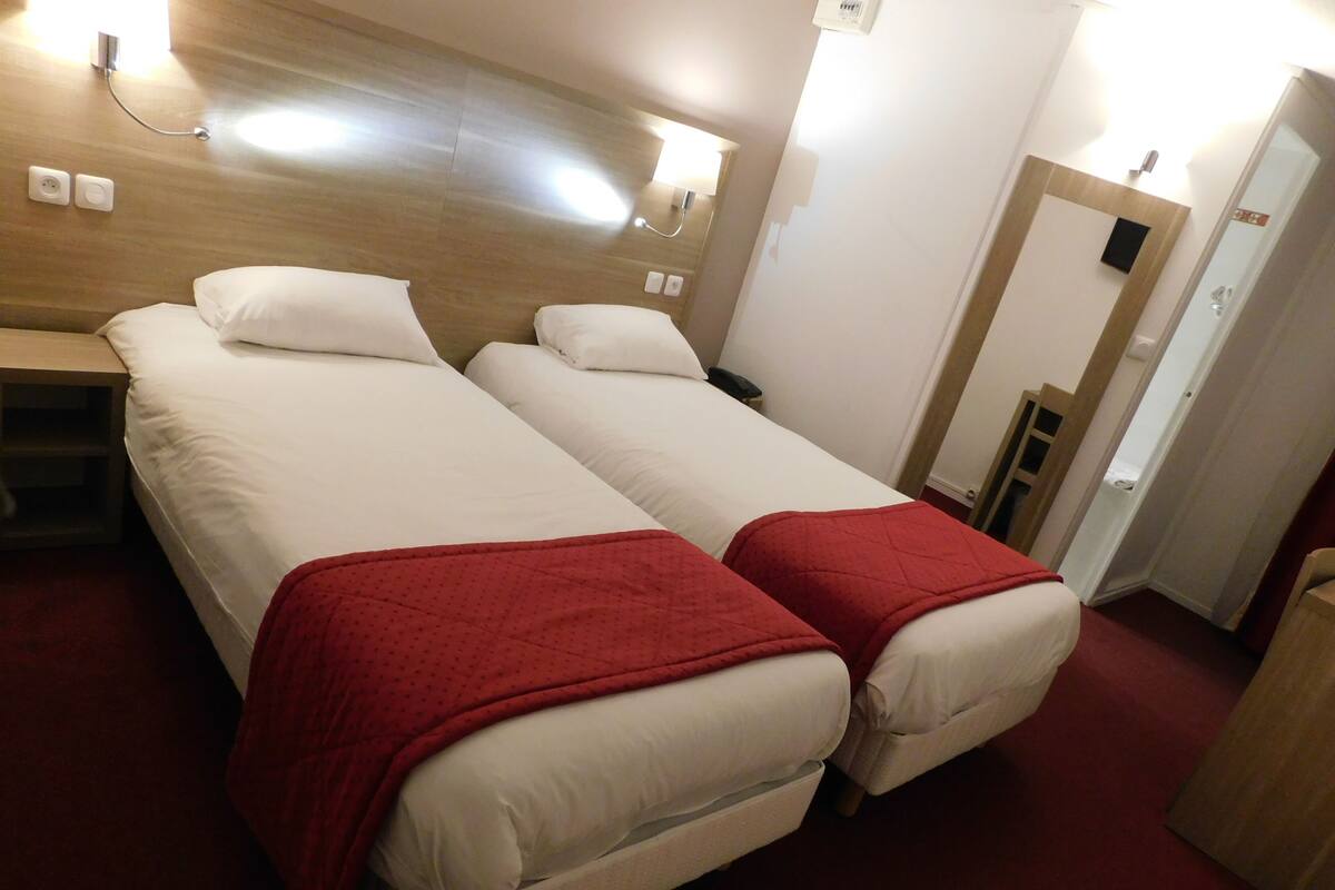 A hotel room with two single beds, each with a red blanket, a wooden headboard, and a bedside table.