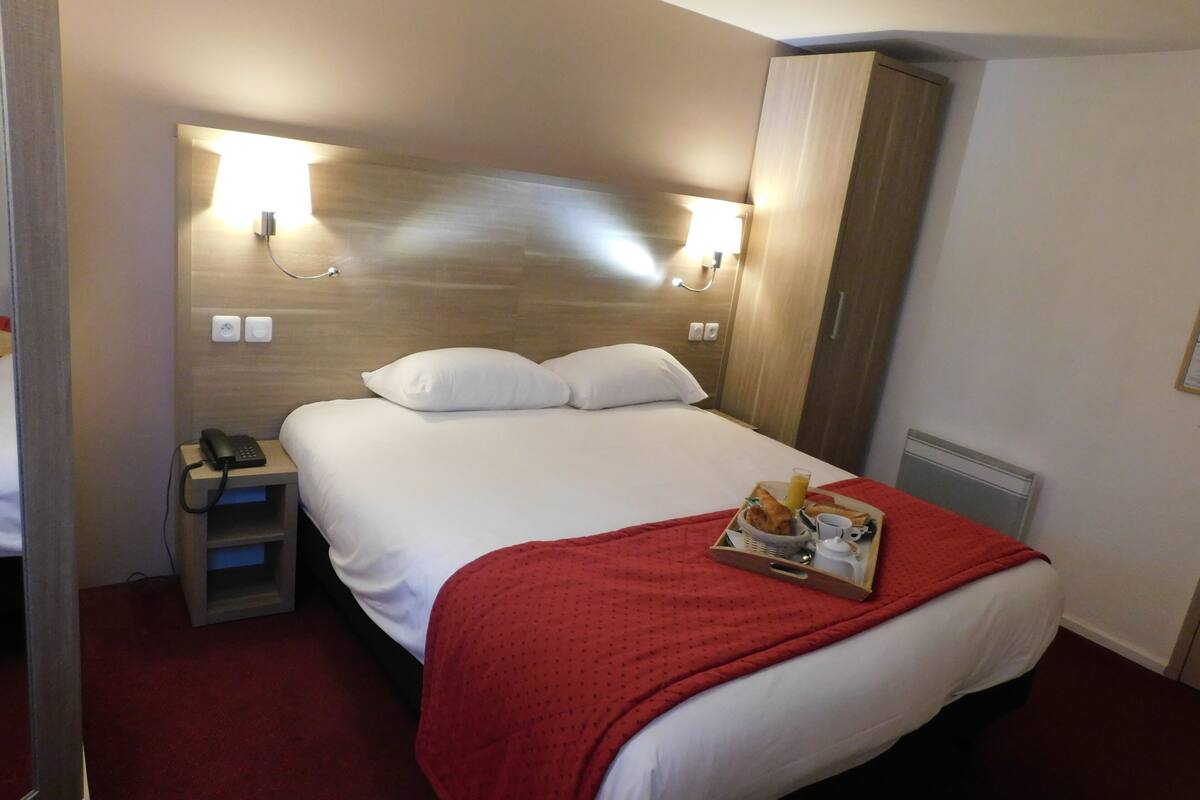 A hotel room with a neatly made bed, a bedside phone, a tray with breakfast items, and a red throw blanket.
