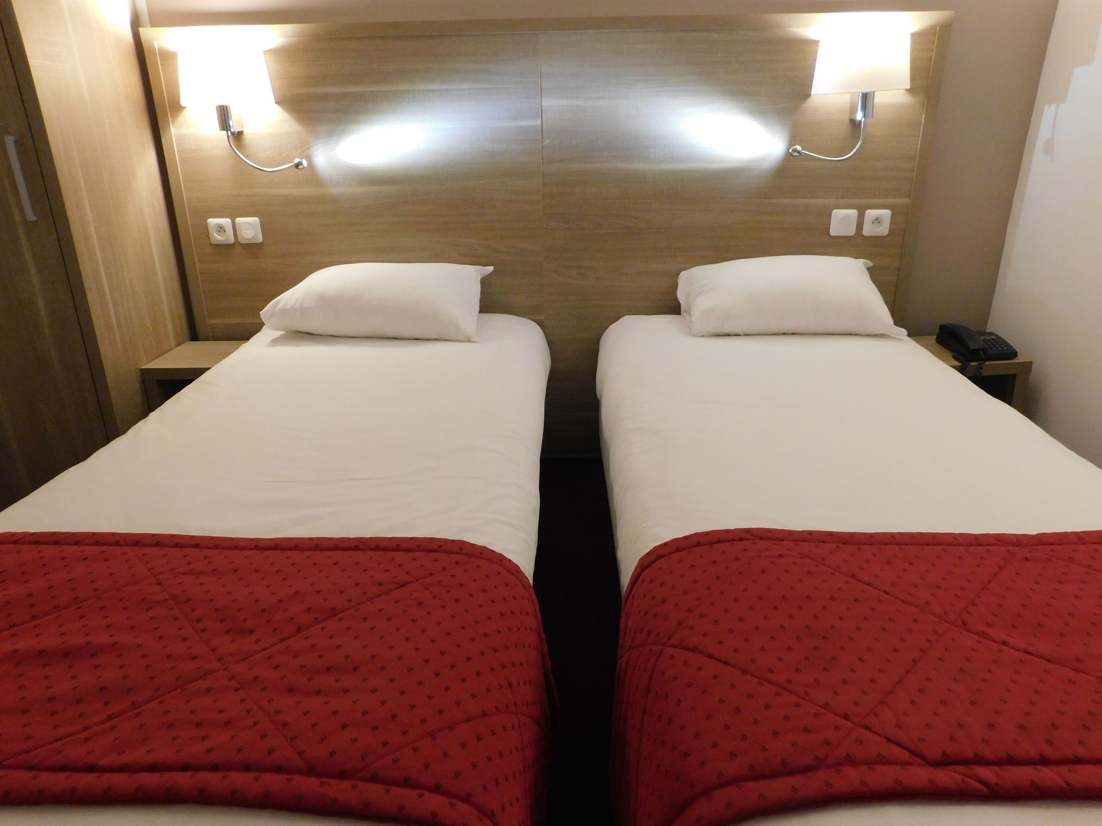 A hotel room with two single beds, each with a white pillow and a red blanket. There are bedside tables with lamps and a phone on one of them.