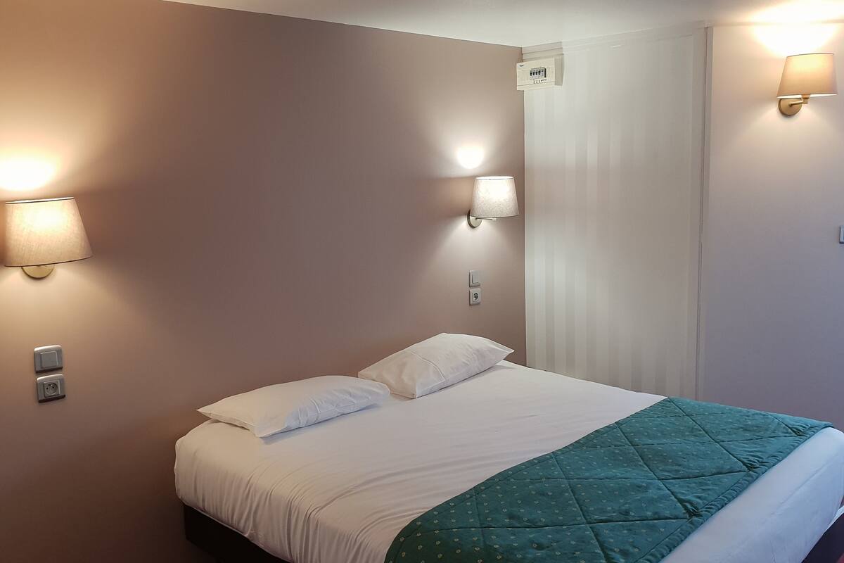 A hotel room with a bed, two wall-mounted light fixtures, a thermostat, and a door with a window.