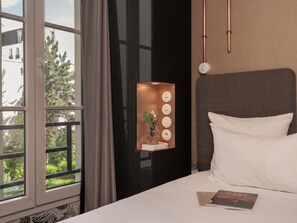 Quarto de hotel com uma janela grande, mesa de cabeceira com um livro, luminária na parede e cabeceira estampada.