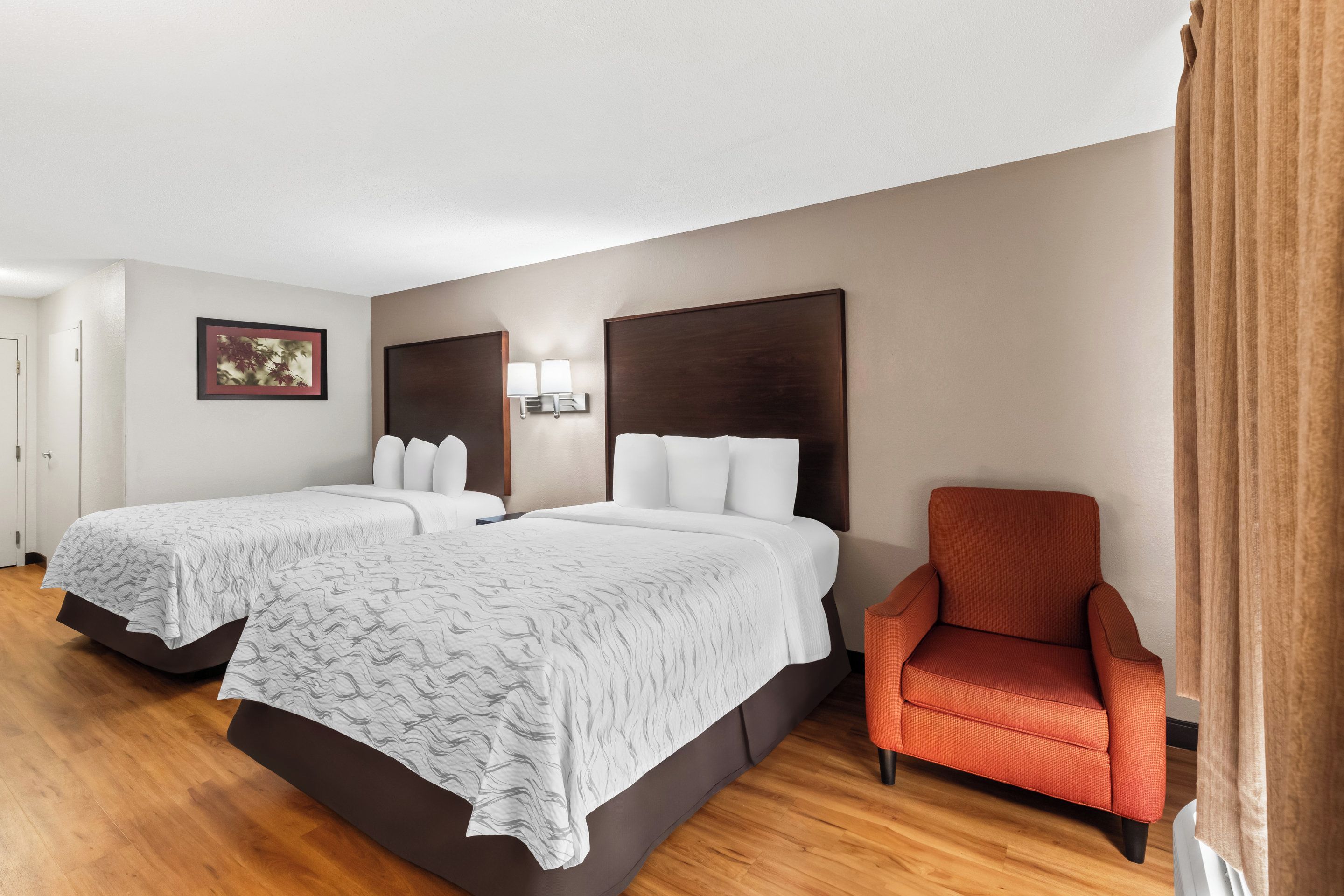 A hotel room with two beds, a chair, and a wall-mounted lamp.