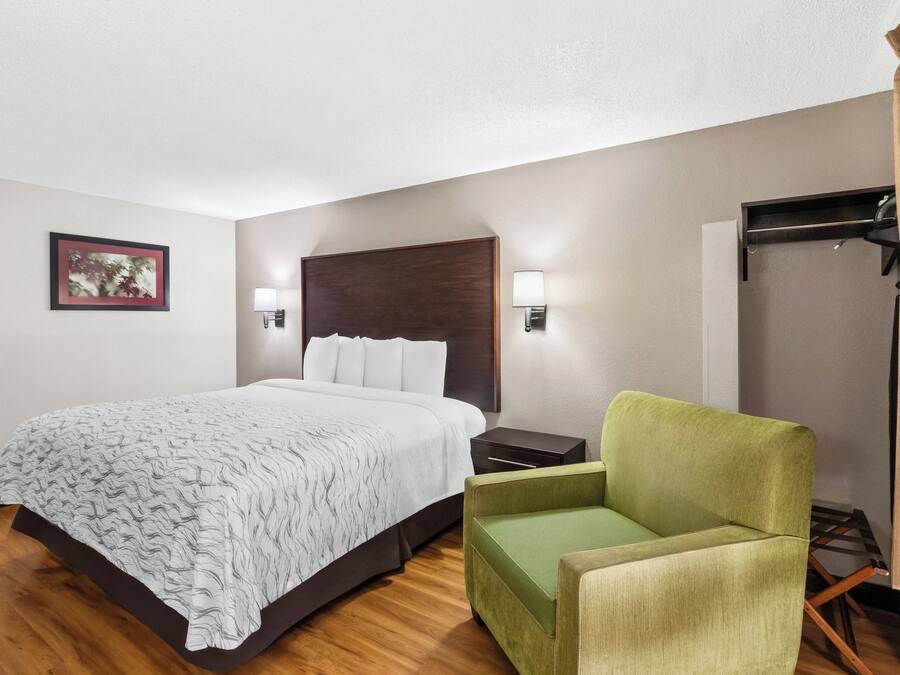 A hotel room with a bed, a green armchair, a nightstand, a wall-mounted lamp, and a framed picture on the wall.