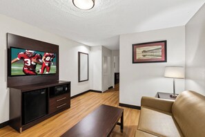 Habitación de hotel con televisión de pantalla plana montada en la pared, una mesa de centro de madera, un sillón color beige y un cuadro enmarcado en la pared.