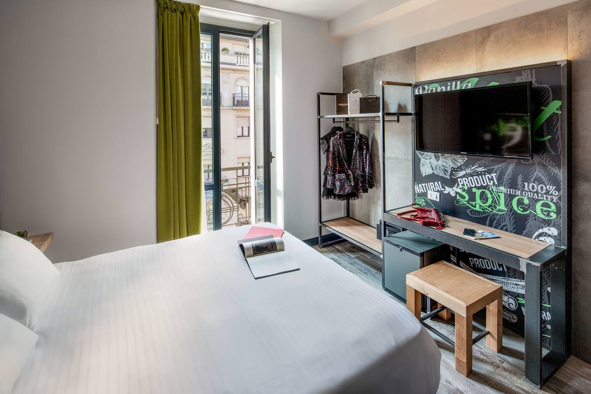 Una camera d'albergo con un letto, una scrivania con un computer, una televisione e una finestra vista edifici.