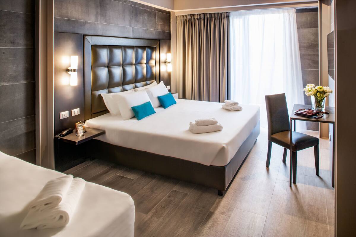 Camera d'albergo con un letto grande, un comodino con una lampada, una sedia e una scrivania con un vaso di fiori.