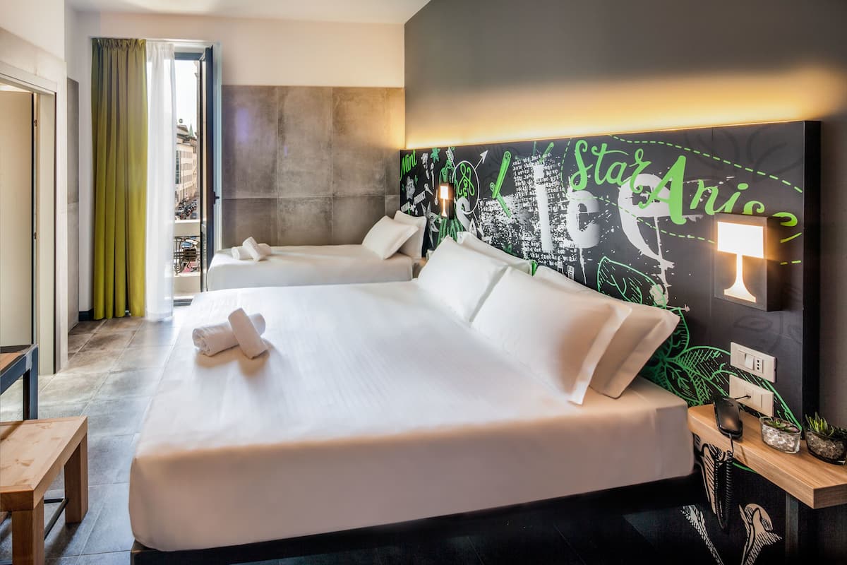 Camera d'albergo con due letti, un grande murale sulla parete, un comodino con una lampada e vista sul balcone esterno.