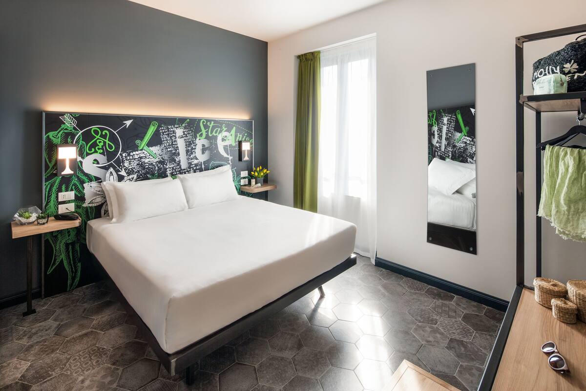A modern hotel room with a large bed, a cityscape artwork on the headboard, a mirror, and a wooden side table with a lamp.