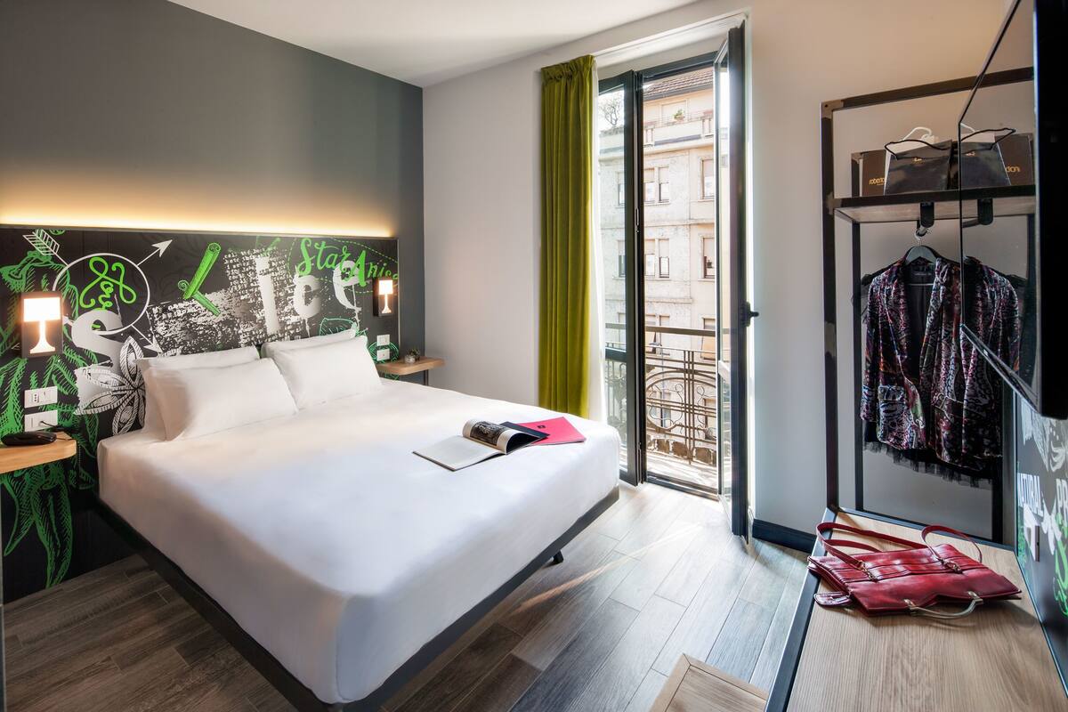 Una camera d'albergo moderna con un letto grande, un comodino, un quadro appeso alla parete, una finestra con vista sugli edifici e un appendiabiti con borse.