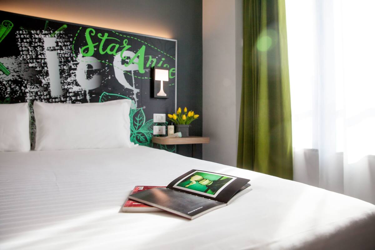 A neatly made bed with a magazine on it, a wall with a green and white design, and a window with curtains.