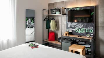 A hotel room with a bed, a desk, a TV, a shelf with a 'Vanilla' sign, and a coat rack with clothes.