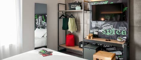 A hotel room with a bed, a desk, a TV, a shelf with a 'Vanilla' sign, and a coat rack with clothes.