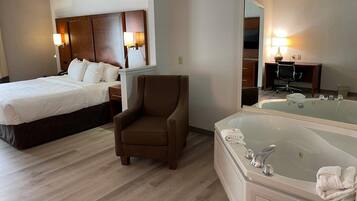 A hotel room with a bed, a brown armchair, a large mirror, a desk with a chair, and a bathtub with towels.