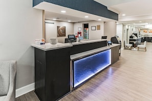A reception desk with a digital screen, a glass partition, and a seating area with chairs and a table.