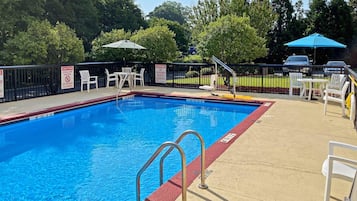 A swimming pool with a red and blue border, surrounded by white chairs and tables, and a black fence with warning signs.