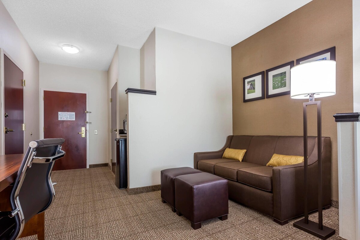 A hotel room with a brown sofa, a matching ottoman, a desk with a chair, a standing lamp, and framed pictures on the wall.