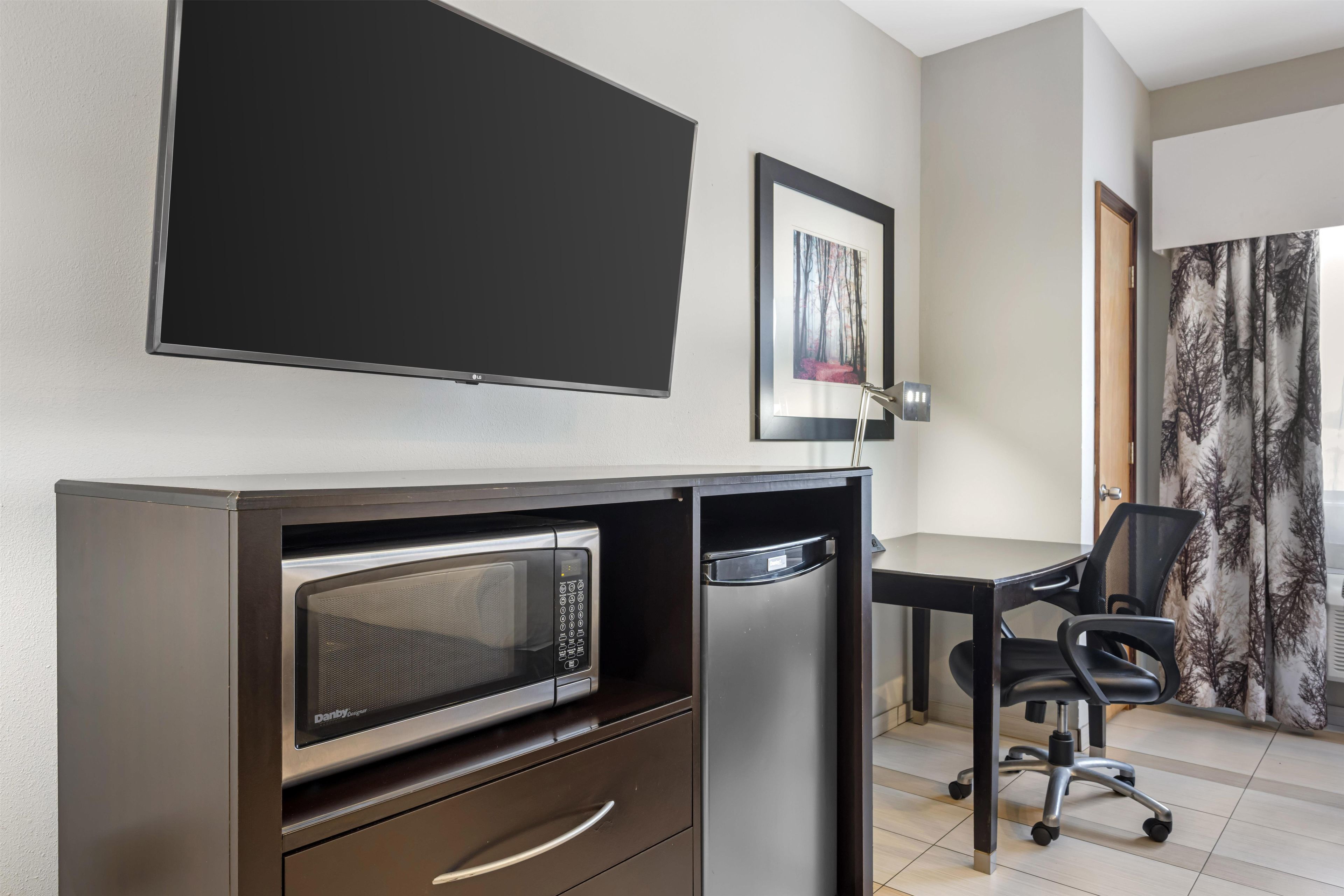 A compact hotel room with a flat-screen TV, a microwave, a small refrigerator, a desk with a chair, and a framed picture on the wall.