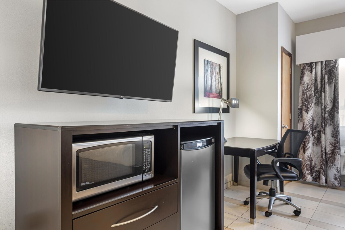 A compact hotel room with a flat-screen TV, a microwave, a small refrigerator, a desk with a chair, and a framed picture on the wall.