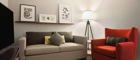 Een moderne woonkamer met een beige bank, een rode fauteuil en een vloerlamp. Er hangen afbeeldingen in lijsten aan de muur en staat een flatscreen televisie op een dressoir.