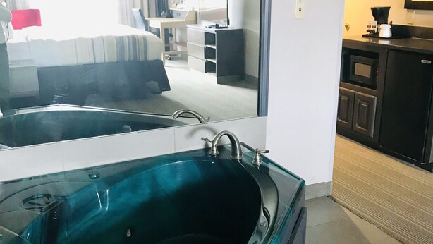 Ein modernes Hotelzimmer mit einer großzügigen, gläsernen Badewanne, einem Bett mit grau-weiß gestreifter Bettdecke, einem Schreibtisch mit Computer und einer Küchenzeile inklusive Kaffeemaschine.