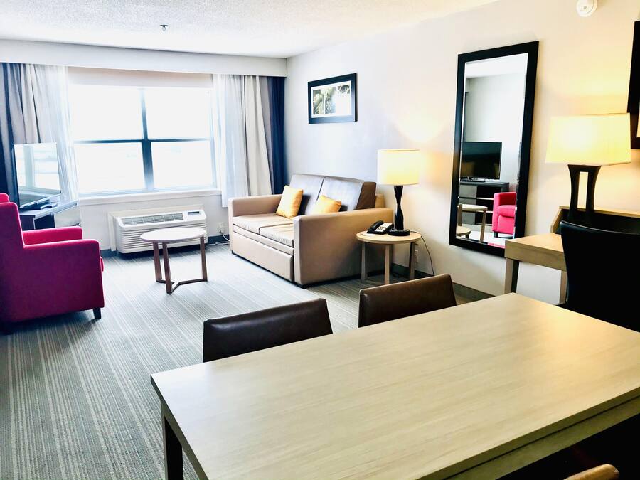 Habitación de hotel con sofá, mesa de comedor, sillón rojo, lámpara y ventana grande.