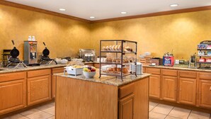 A hotel kitchen with a self-service breakfast station, including waffles, pastries, and a fruit bowl.