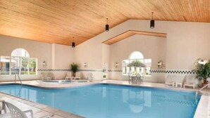 An indoor swimming pool with a wooden ceiling, tiled floor, and a poolside area with chairs and tables.