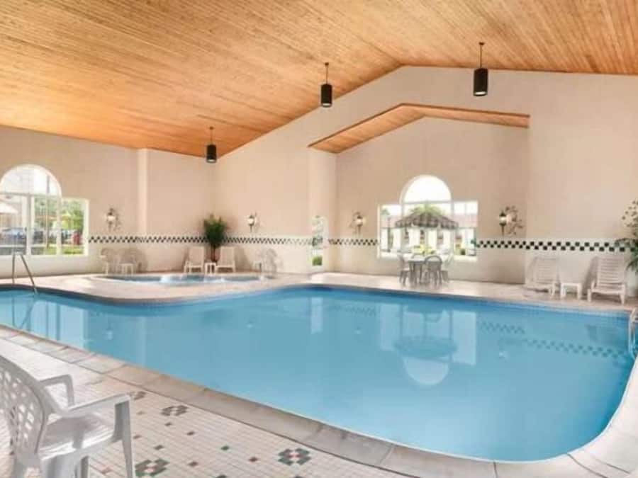 An indoor swimming pool with a wooden ceiling, tiled floor, and a poolside area with chairs and tables.