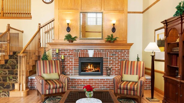 Una sala de estar con chimenea, dos sillones a rayas, una mesa de centro con un jarrón de flores, una escalera y un armario de madera.