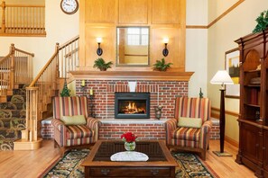 A living room with a fireplace, two striped armchairs, a coffee table with a vase of flowers, a staircase, and a wooden cabinet. - Country Inn & Suites by Radisson, Sycamore, IL (Sycamore)
