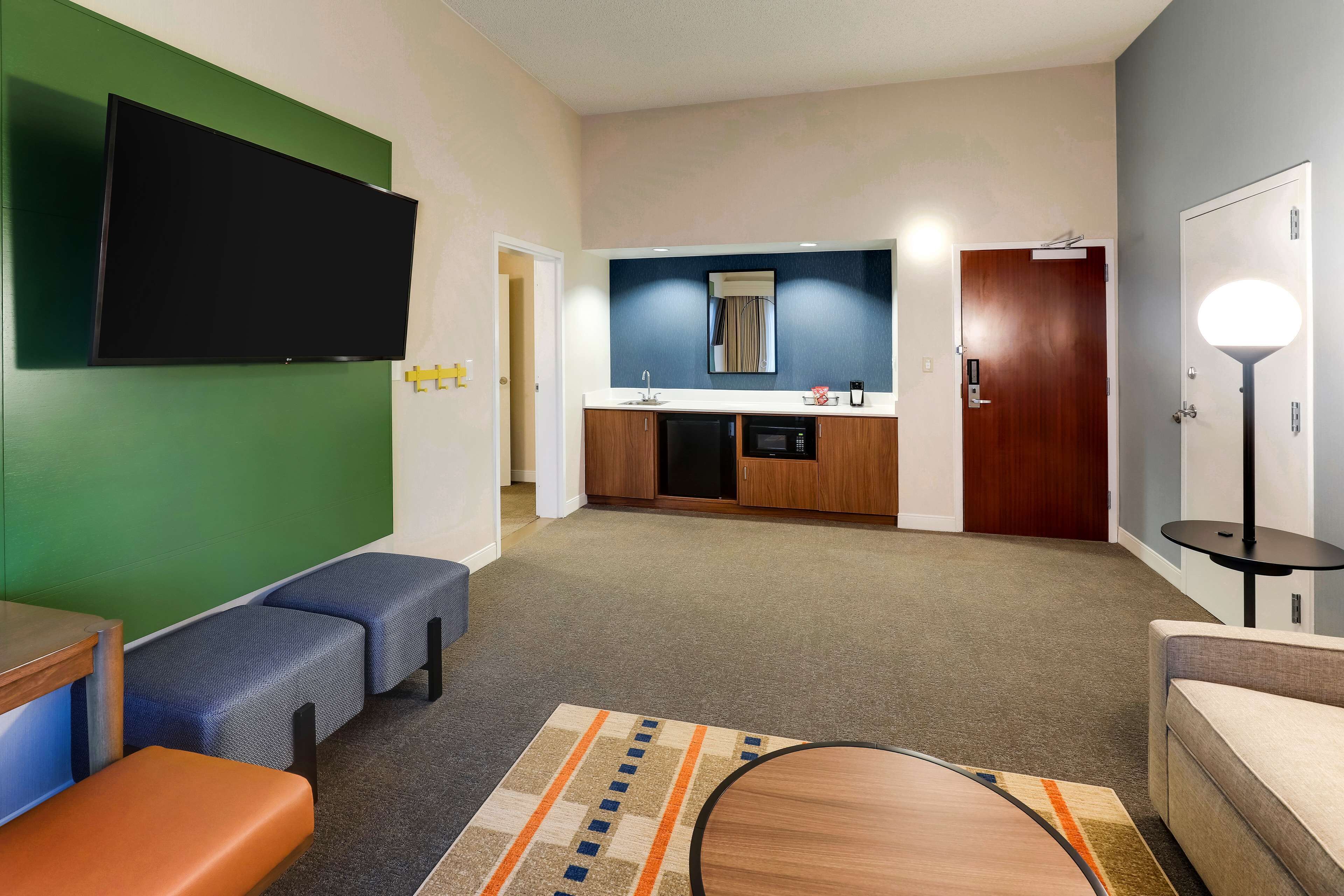 Una habitación de hotel moderna con televisor de pantalla plana, kitchenette, zona de estar con sofá y sillones, y una pequeña mesa redonda.