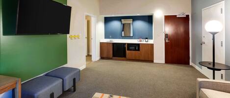 Una habitación de hotel moderna con televisor de pantalla plana, kitchenette, zona de estar con sofá y sillones, y una pequeña mesa redonda.