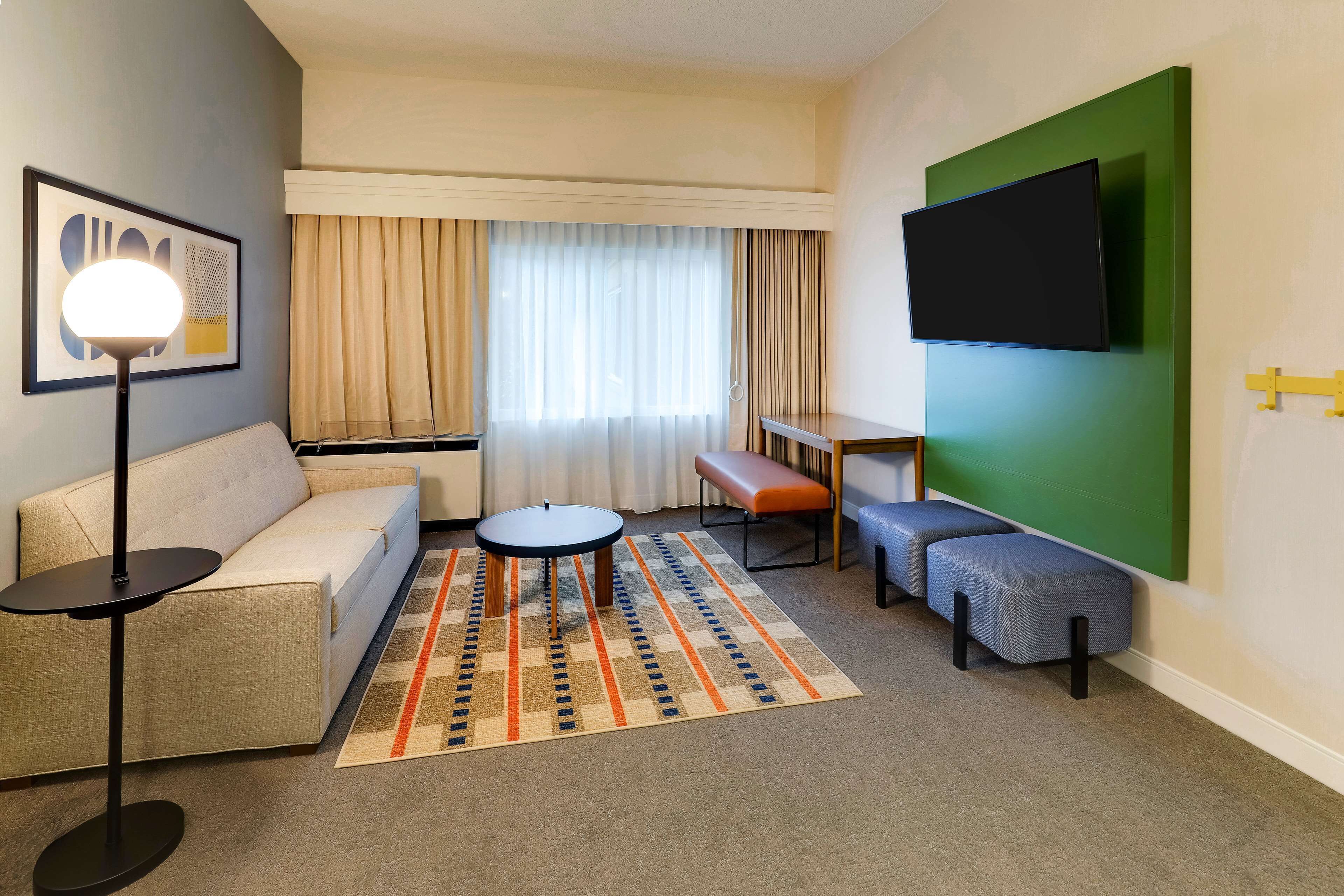 Moderni hotellihuone, jossa on sohva, pyöreä sohvapöytä, penkki ja seinälle asennettu litteänäyttöinen televisio. Seinä on maalattu vihreäksi.