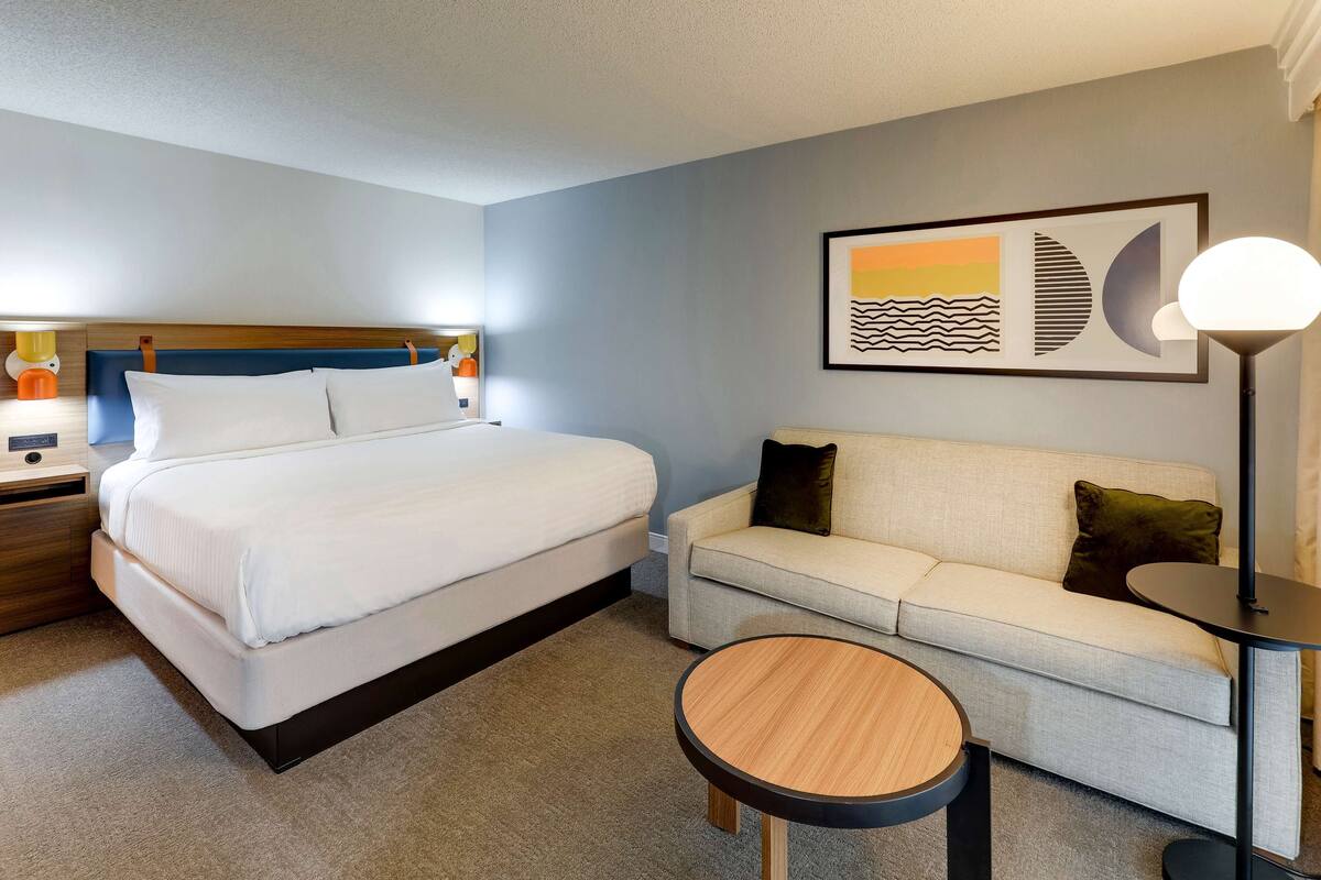 Un cuarto de hotel con cama, sofá, una mesa redonda de madera, lámpara y un cuadro abstracto enmarcado en la pared.