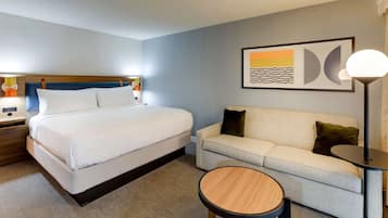 A hotel room with a bed, a sofa, a round wooden table, a lamp, and a framed abstract painting on the wall.