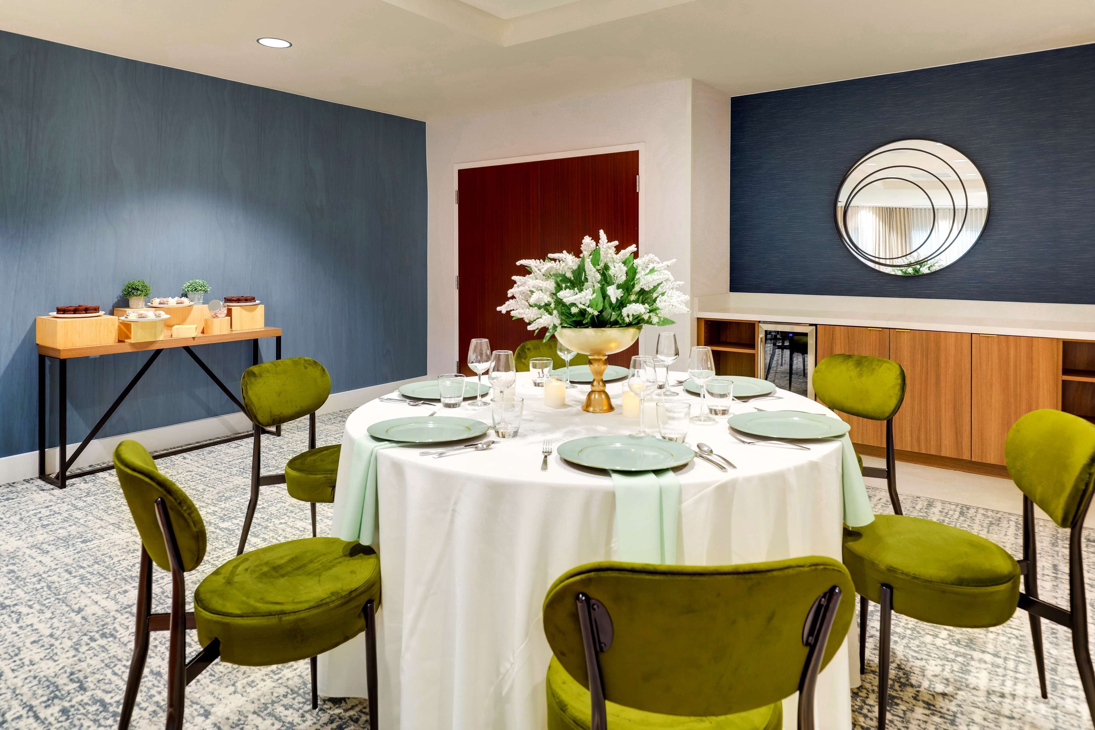 A dining area with a round table set for six, surrounded by green upholstered chairs. A buffet table with various dishes is visible in the background.