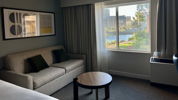 A hotel room with a sofa, a small round table, a large window with a view of buildings and a body of water. - Sonesta Select San Francisco Airport Oyster Point Waterfront (South San Francisco)