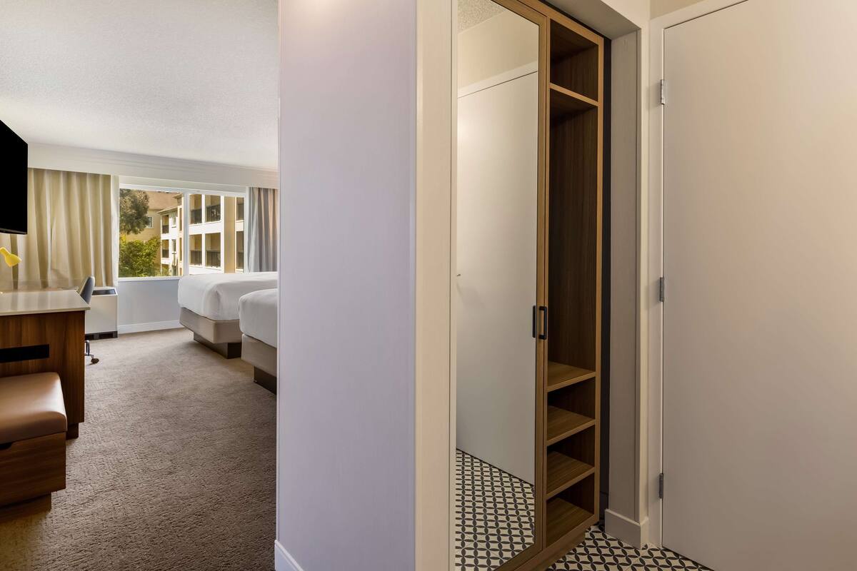Una habitación de hotel con cama, escritorio, silla, armario y ventana con cortinas.