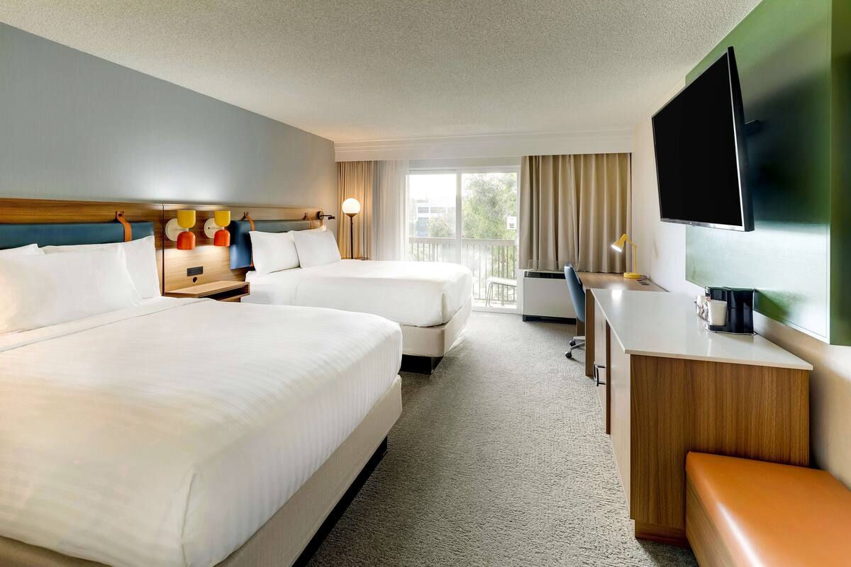 Habitación de hotel con dos camas, un escritorio, un televisor y un balcón con cortinas.