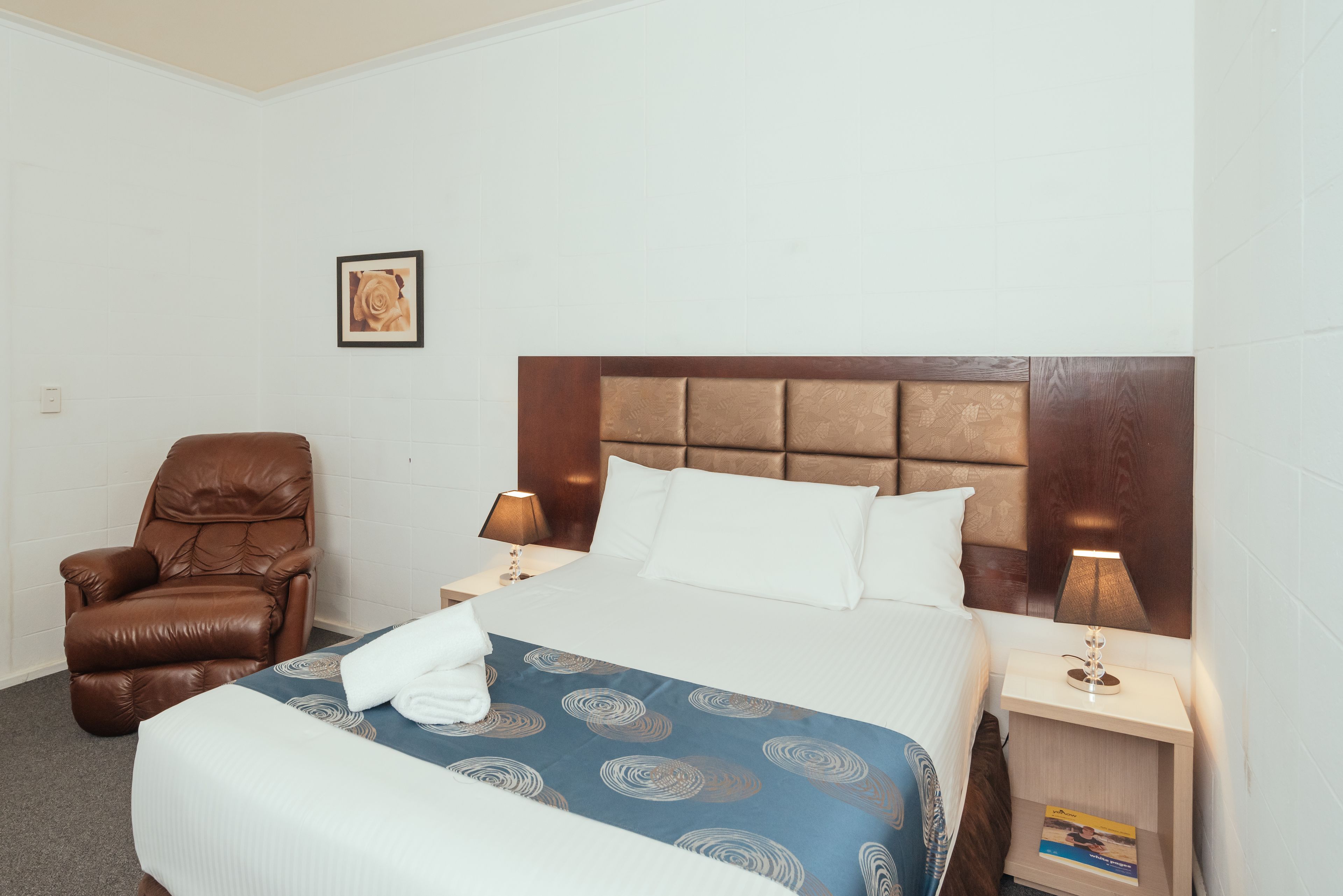 A hotel room with a large bed, two bedside tables, a brown leather chair, a framed picture on the wall, and a blue patterned bedspread.