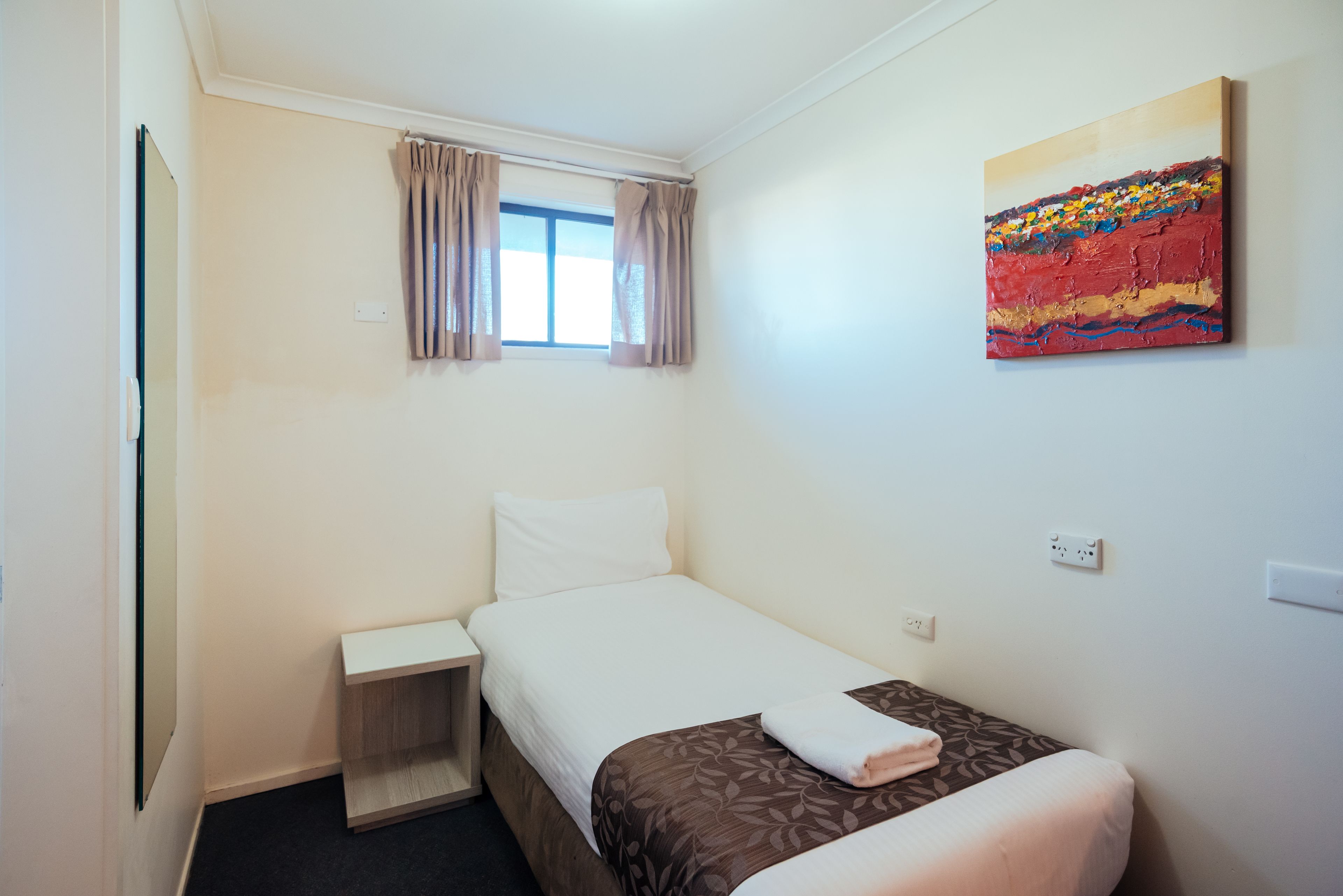 A small, neatly arranged bedroom with a single bed, a bedside table, a window with curtains, and a wall-mounted painting.
