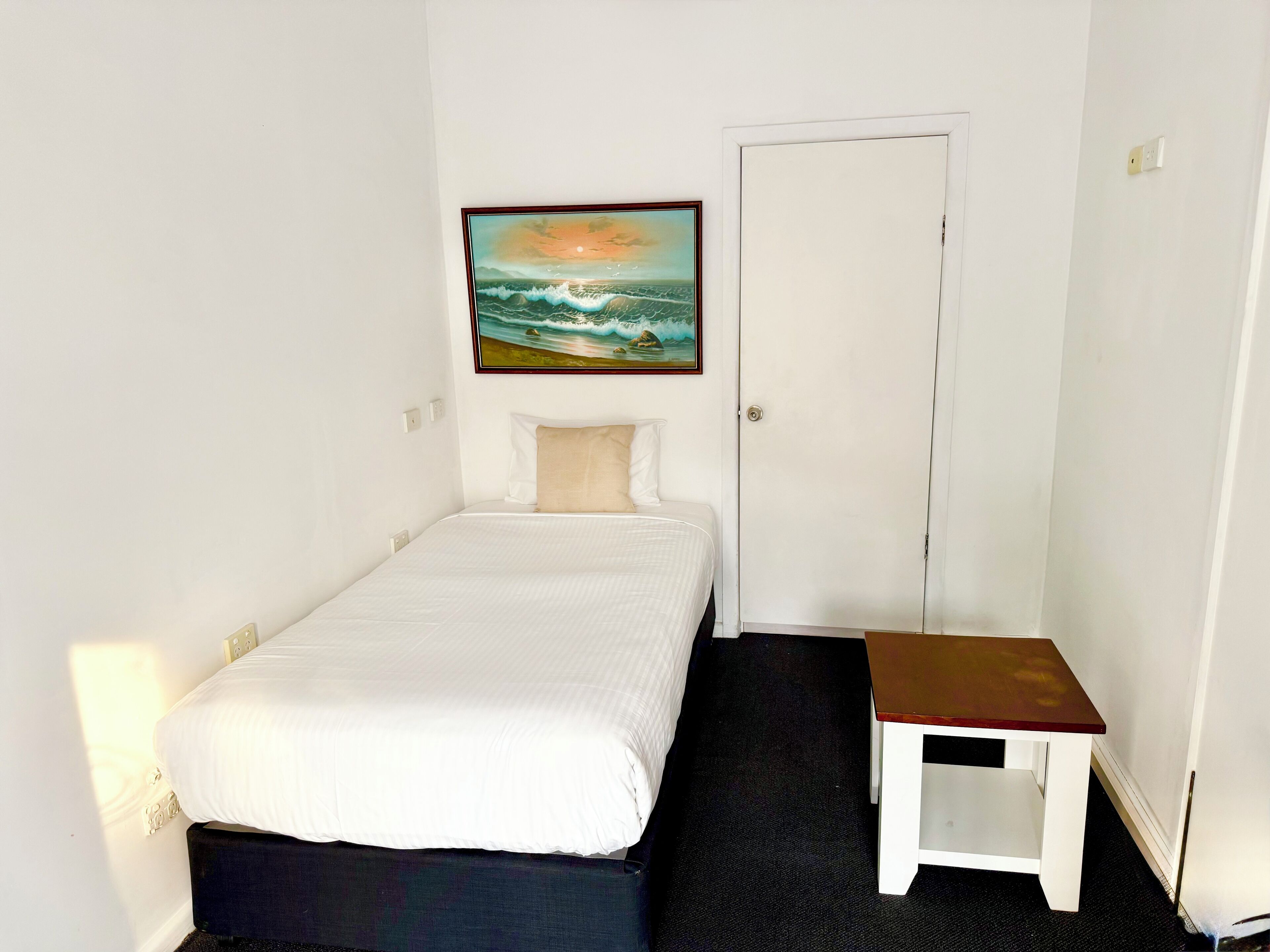 Une petite chambre avec un lit simple, une petite table et un tableau représentant un coucher de soleil sur l’océan, accroché au mur.