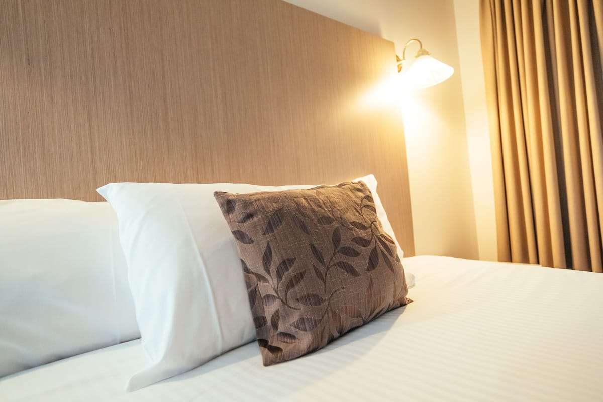A neatly made bed with white and brown pillows, a wooden headboard, and a wall-mounted light fixture.