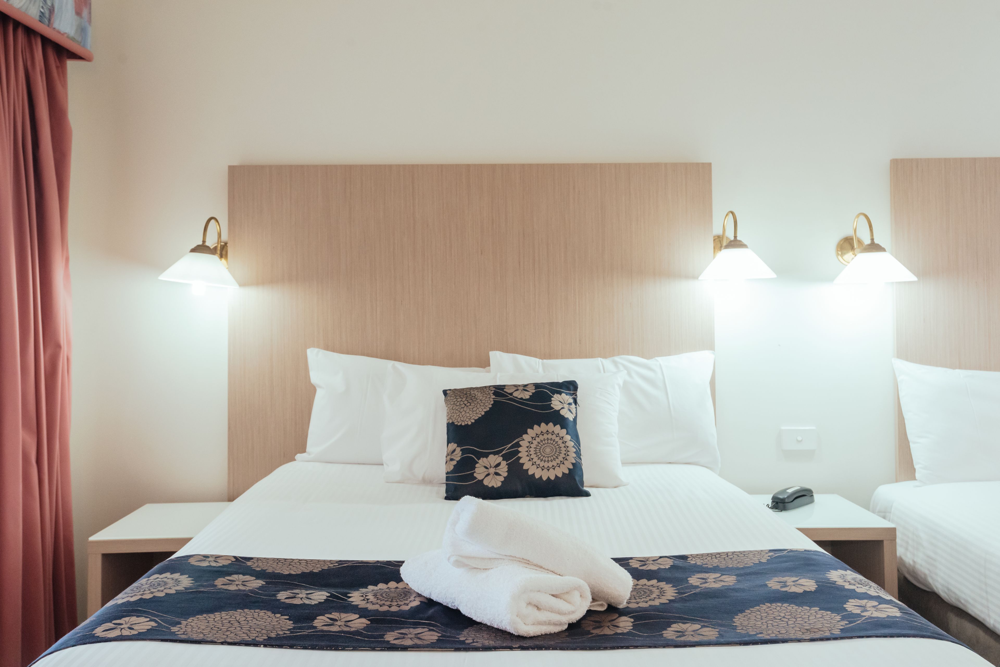 A hotel room with a bed, two bedside tables, a nightstand, a decorative pillow, and wall-mounted lights.